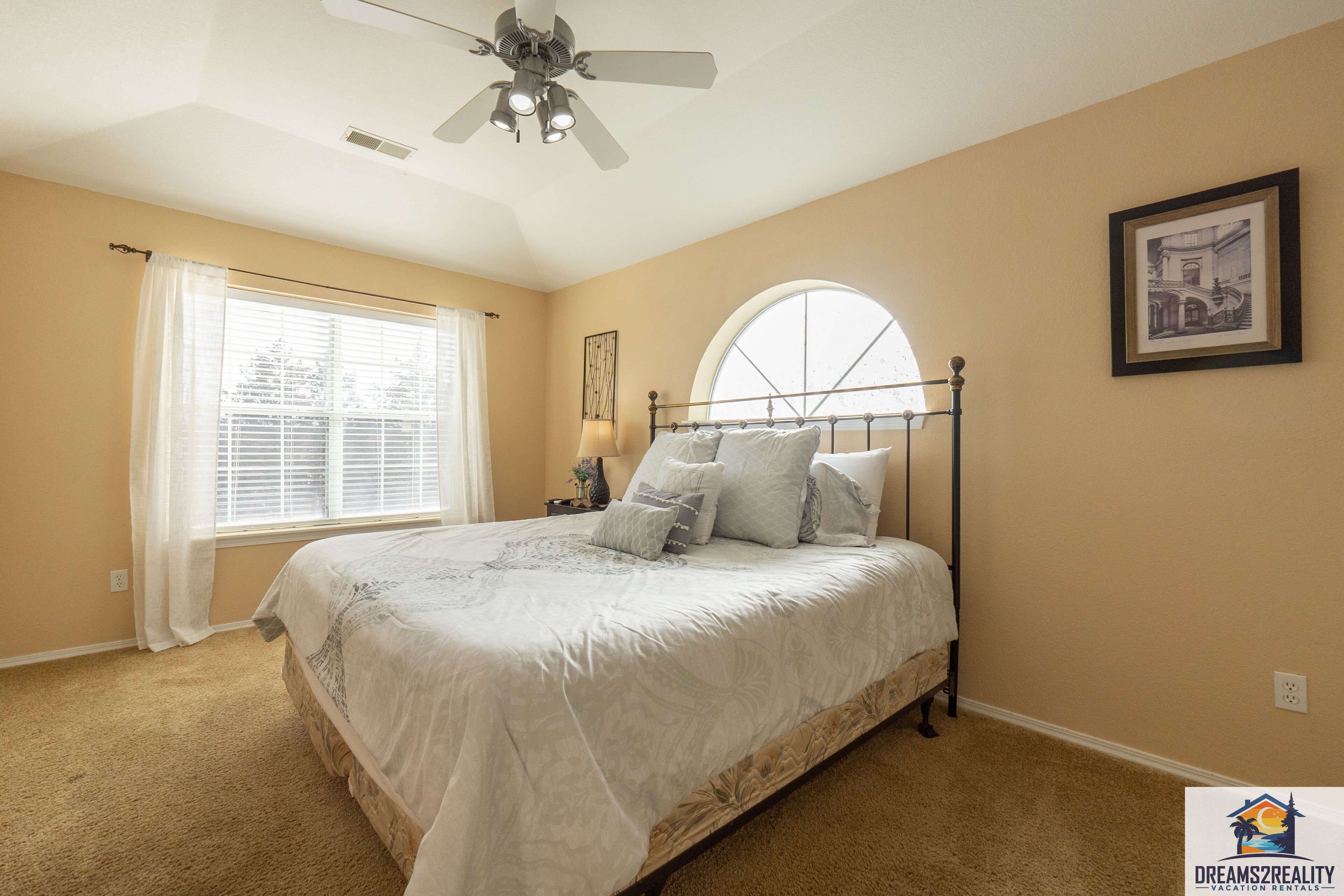 A bedroom with a bed, a ceiling fan, a window with blinds, a lamp, and a framed picture on the wall.
