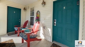A porch with two red Adirondack chairs, a teal door, and a small window with a flower arrangement.