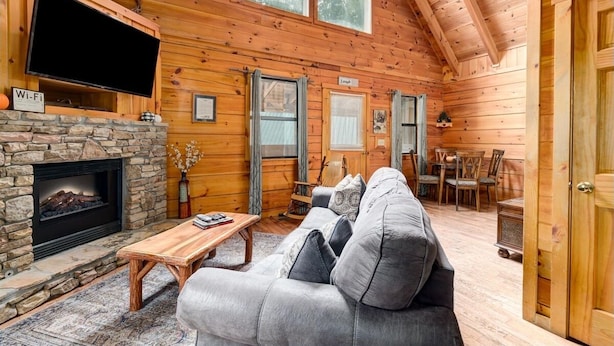 A cozy cabin interior with a stone fireplace, a flat-screen TV mounted above it, a grey sofa, and a dining area with wooden chairs and a table.
