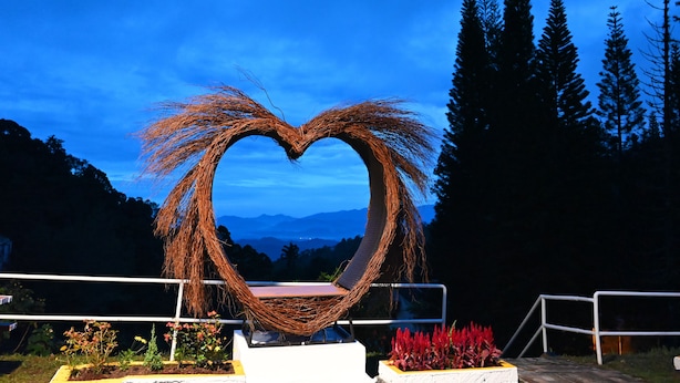 A heart-shaped wicker sculpture with a bench in front, surrounded by greenery and mountains in the background. - The Pines (Raub)