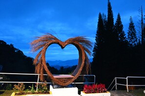 Escultura de mimbre con forma de corazón y un banco en primer plano, rodeada de vegetación y montañas al fondo.