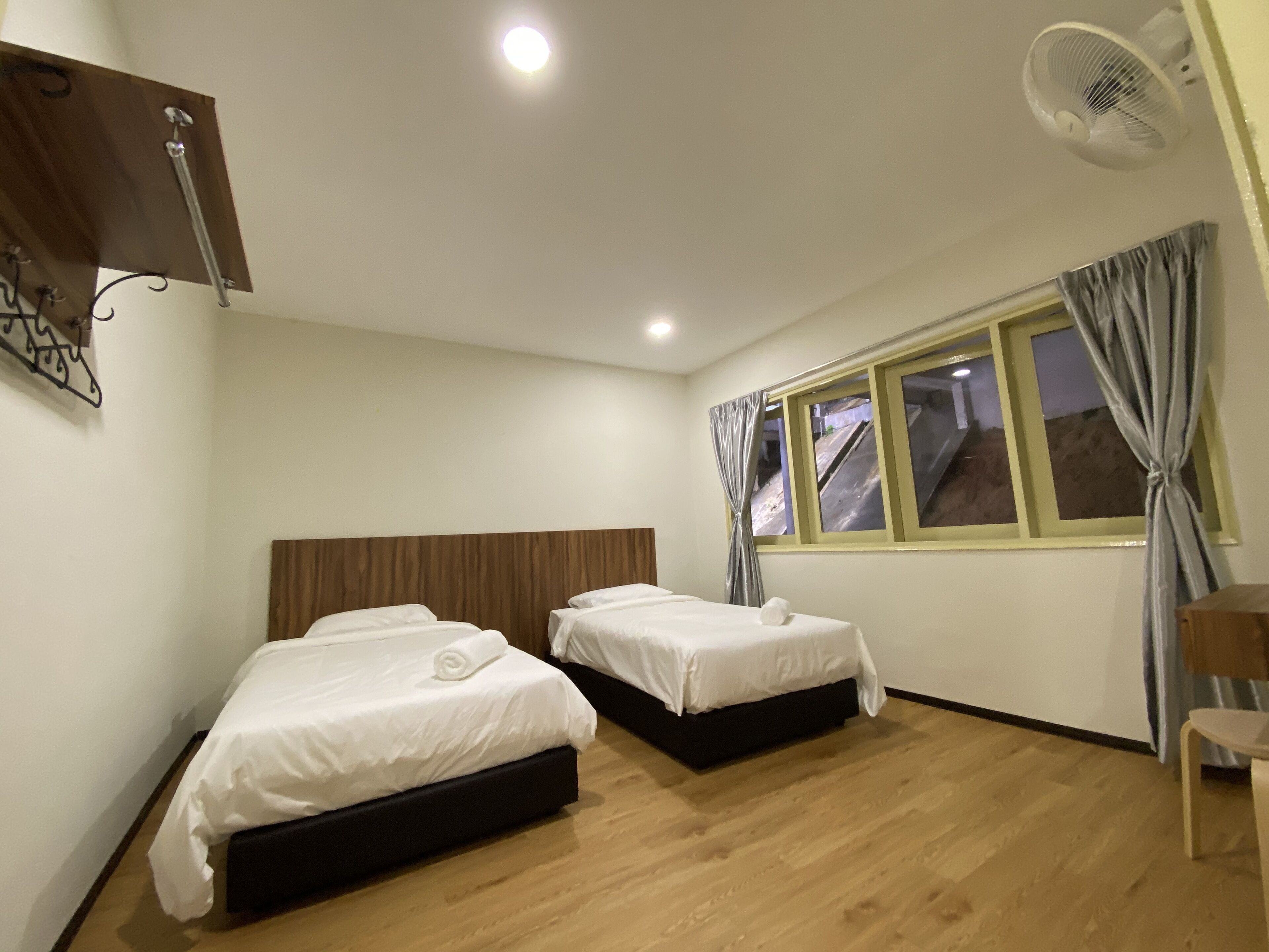A hotel room with two beds, a wooden floor, a window with curtains, a ceiling fan, and a wall-mounted light fixture.