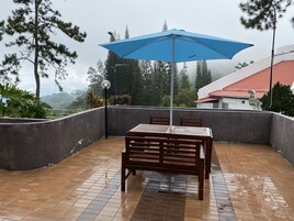 Um alpendre coberto com mesa e cadeiras, um guarda-sol azul e vista para árvores e edifícios ao fundo.