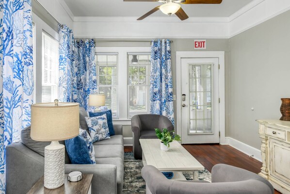 A living room with a sofa, armchair, coffee table, and a lamp. There is a door with an exit sign, a ceiling fan, and a window with blinds.