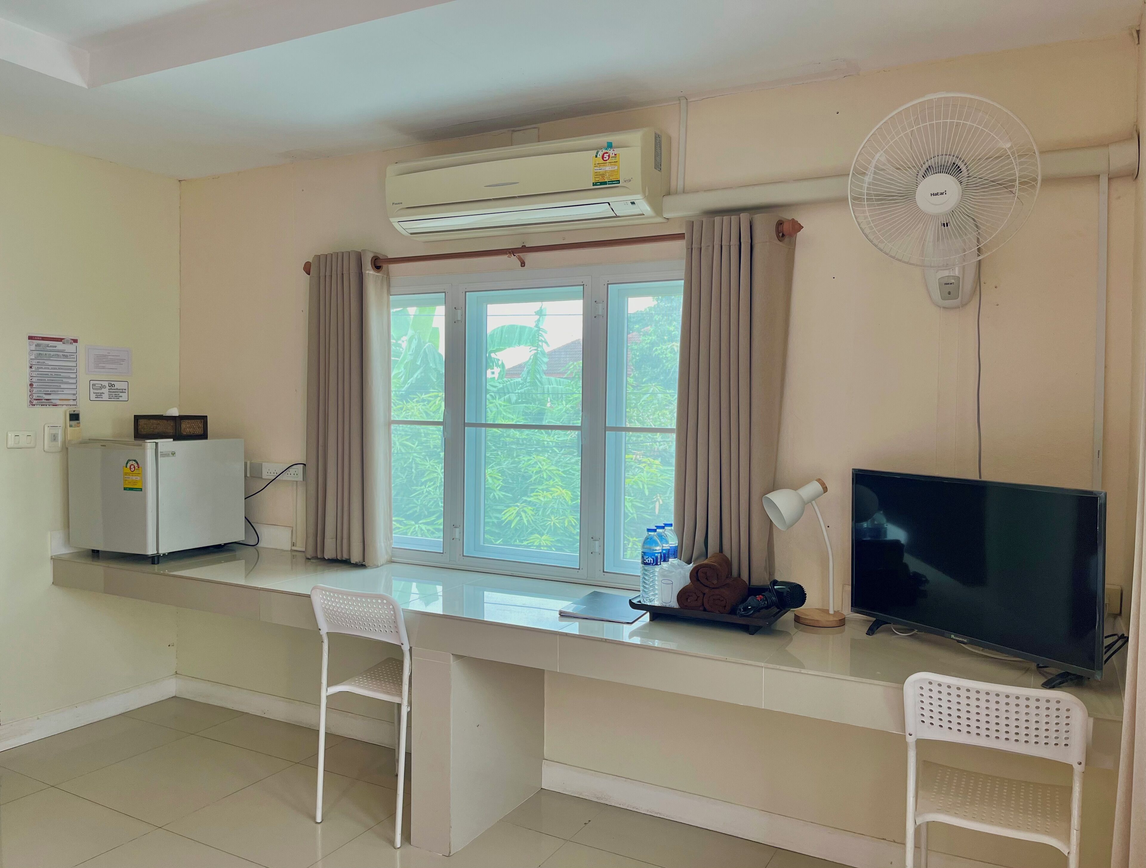 A hotel room with a desk, two chairs, a television, an air conditioner, a fan, a window with curtains, and a small refrigerator.