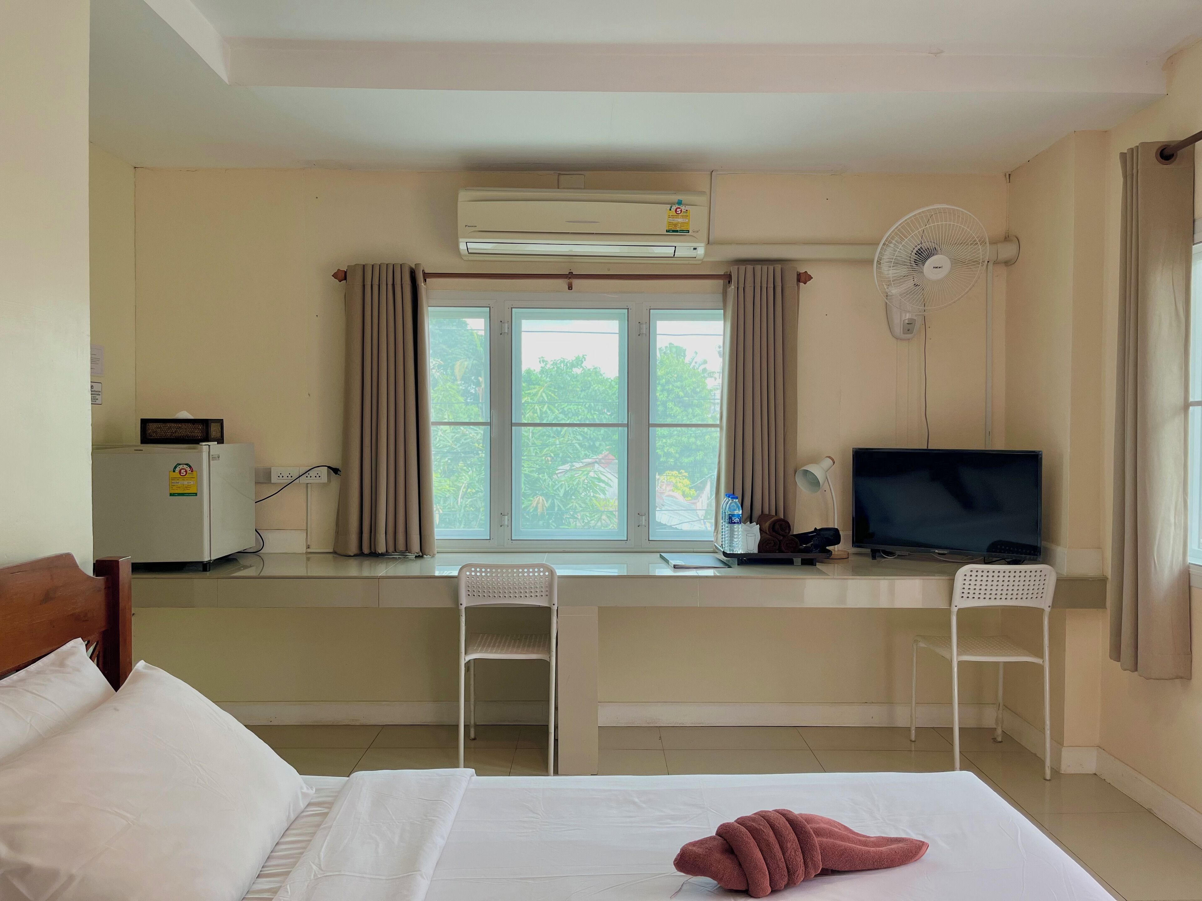 A hotel room with a bed, a desk with a chair, a TV, an air conditioner, and a window with curtains.