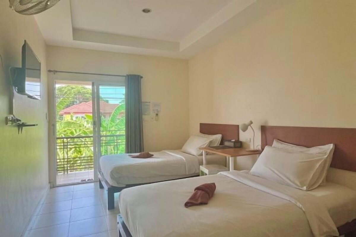 A hotel room with two beds, a small table, a nightstand, and a view of greenery through the window.
