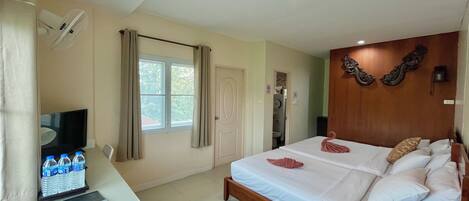A hotel room with a bed, a desk with a laptop and water bottles, a TV, a window with curtains, and a bathroom door.