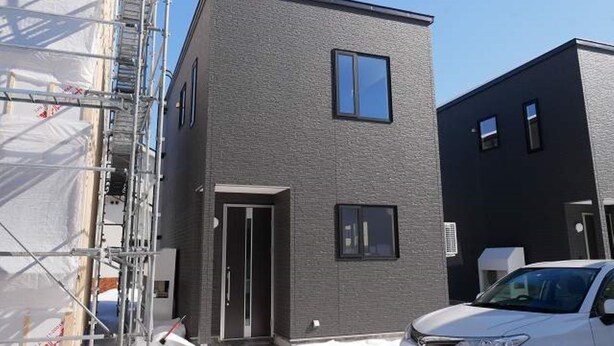 A modern two-story building with a dark gray facade, a visible entrance, and a parked white car. - Nagayama Whole Apartment (Asahikawa)
