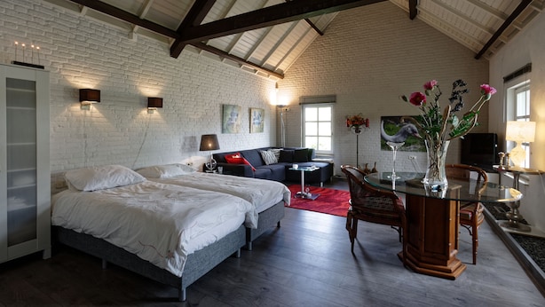 A spacious room with a high ceiling, exposed brick walls, and a large bed with two pillows. There is a glass table with a vase of flowers and a chair. - Room-room to Rent With own Entrance, liv (Blitterswijck)