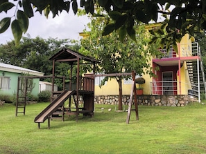 A playground with a slide and swing set, surrounded by grass and trees. - Cabaña del Sol Ground Floor 2 Bedroom Cabin for 6 Guests (Vaqueros)