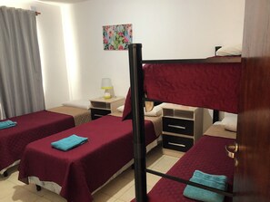 A room with two beds, each with a red bedspread and a blue towel. There is a nightstand with a lamp, a small table with a lamp, and a wall-mounted painting.