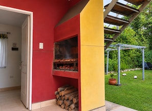 Uma área de churrasco ao ar livre com churrasqueira vermelha e amarela, uma pilha de lenha e um jardim com pérgola.