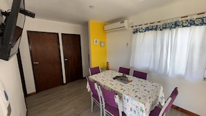 Uma área de refeições com mesa, cadeiras, televisão e uma janela com cortinas.