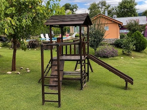 A wooden play structure with a slide and a ladder, surrounded by grass and trees.