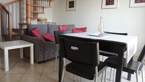 Un salon moderne avec un canapé gris, des coussins rouges, une table basse blanche et une table à manger noire avec des chaises. Un escalier en colimaçon en bois est visible à l’arrière-plan.
