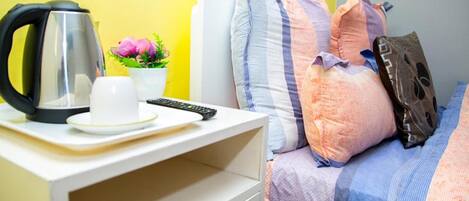 A bed with colorful pillows, a bedside table with a kettle, a cup, and a remote control.