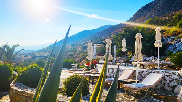 A sunlit outdoor seating area with lounge chairs and umbrellas, surrounded by greenery and a mountainous backdrop.