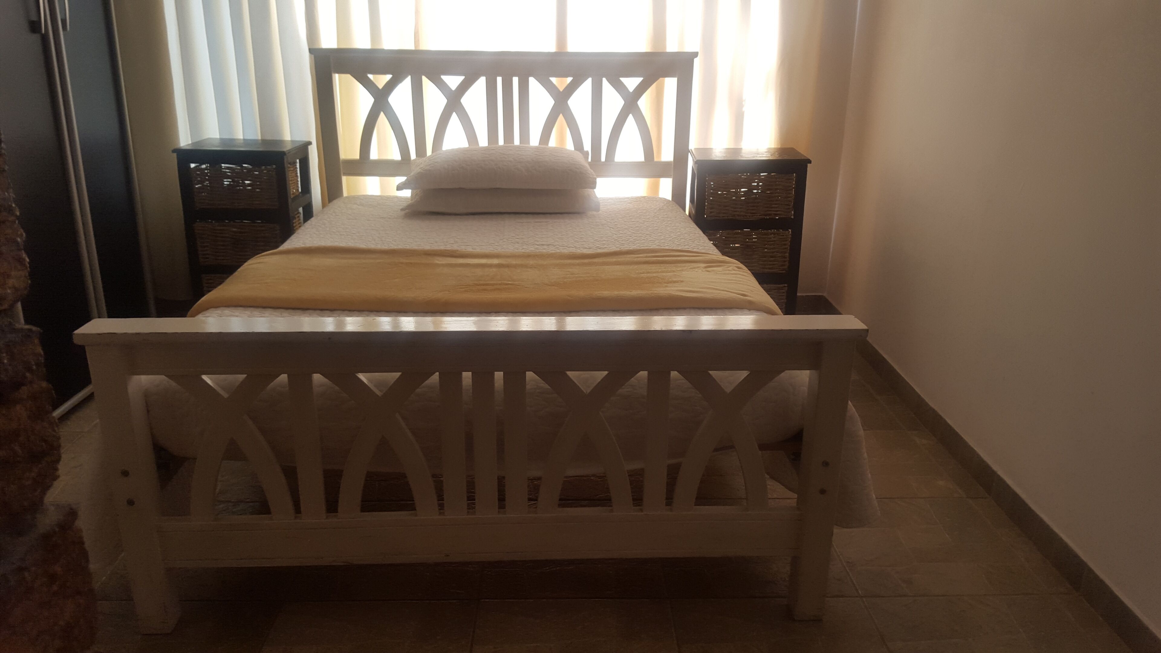 Cama com estrutura de madeira, cabeceira branca e duas mesas de cabeceira com cestos de vime.