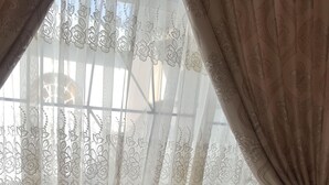 A beige sofa with a cushioned seat and backrest, positioned in front of a window with sheer curtains featuring a floral pattern. - Room in House - Patzerella's Cozy Nest (Krugersdorp)