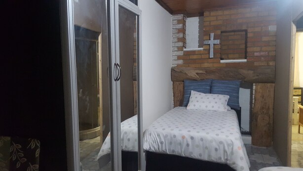 A bedroom with a bed, a wooden headboard, a brick wall, and a cross decoration. - Room in House - Patzerella's Cozy Nest (Krugersdorp)