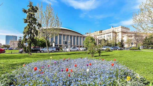 A large building with a curved facade, surrounded by a well-maintained lawn with a variety of flowers and trees. - Amazon Apartment - Cismigiu Gardens for 2 Guests (Bucharest)