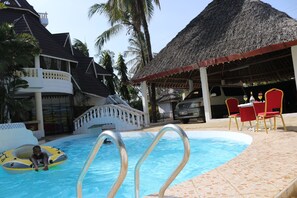 A swimming pool with a thatched-roof structure, a covered seating area, and a poolside chair.
