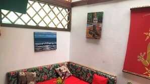 A corner room with a wooden bench, a small table, a wall with a tropical-themed cushion, a framed picture of the ocean, and a red decorative panel.