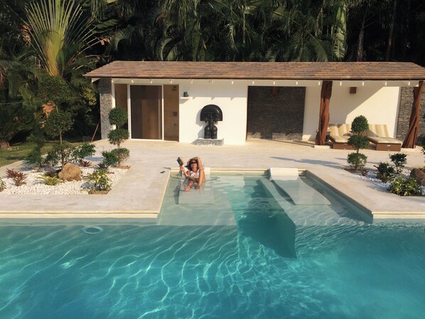 A swimming pool with a person relaxing in the water, surrounded by a landscaped garden with various plants and a building with a tiled roof in the background. - Las Terrenas Front Beach And Garden Villa With Private Staff (Las Terrenas)