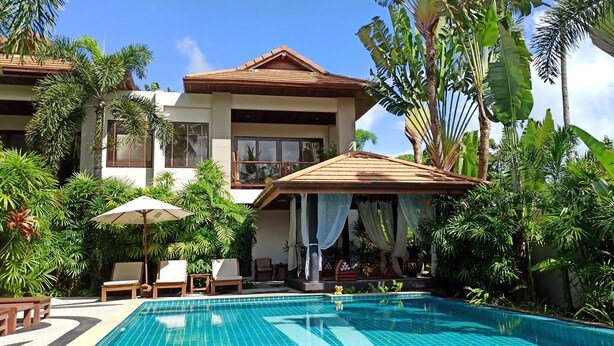 Un espace aménagé au bord de la piscine, avec une piscine carrelée, des chaises longues et un coin salon couvert offrant une vue sur un bâtiment à deux étages entouré de plantes tropicales.