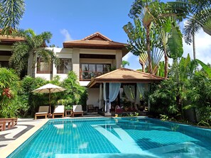 Zona junto a la piscina con una pileta revestida, sillas de descanso y un espacio de estar cubierto con vistas a un edificio de dos pisos rodeado de plantas tropicales.