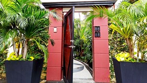 Une porte rouge surmontée d’une enseigne, entourée d’une végétation luxuriante et de plantes en pot.