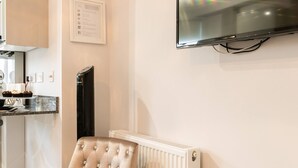 Un salon avec un fauteuil à capitons, une table basse en verre ornée d’un vase de fleurs, un téléviseur à écran plat fixé au mur, et un coin cuisine à l’arrière-plan.
