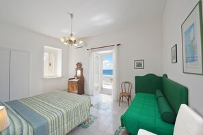 A room with a bed, a green sofa, a wooden dresser, a chair, and a view of the sea through a door. - Villa Panorama Sea View (Furore)