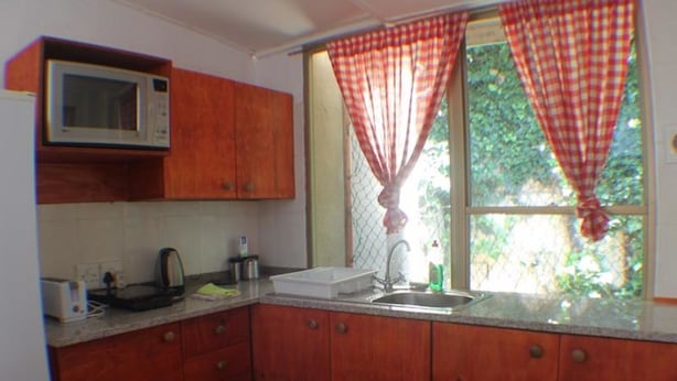 Uma cozinha compacta com armários de madeira, micro-ondas, pia e janela com cortinas vermelhas.