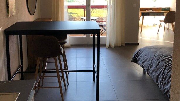 A room with a dining table, two stools, a bed, a hanging lamp, and a view of the outdoors. - Amazing Studio In Lausanne Close To Epfl and Unil (Ecublens)
