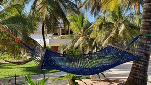 A hammock between palm trees, a building with a white facade, and a clear blue sky. - Mbv Hotel Annex Standard Bungalow (Matemwe)