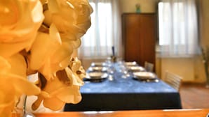 Een gedekte eettafel met borden en bestek, een bloevas en uitzicht op een kamer met raam en gordijnen.