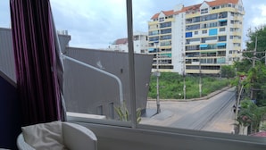 A balcony with a wicker chair and a small table, overlooking a street with buildings and greenery. - 201 Jomtien Hill Resort Sudio (Pattaya)