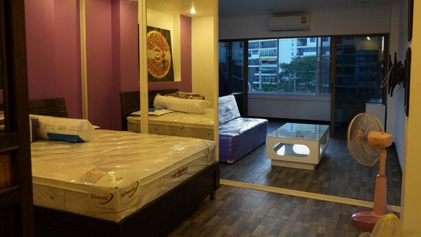A bedroom with a large mirror, a bed with a mattress, a small sofa, and a standing fan. - 201 Jomtien Hill Resort Sudio (Pattaya)