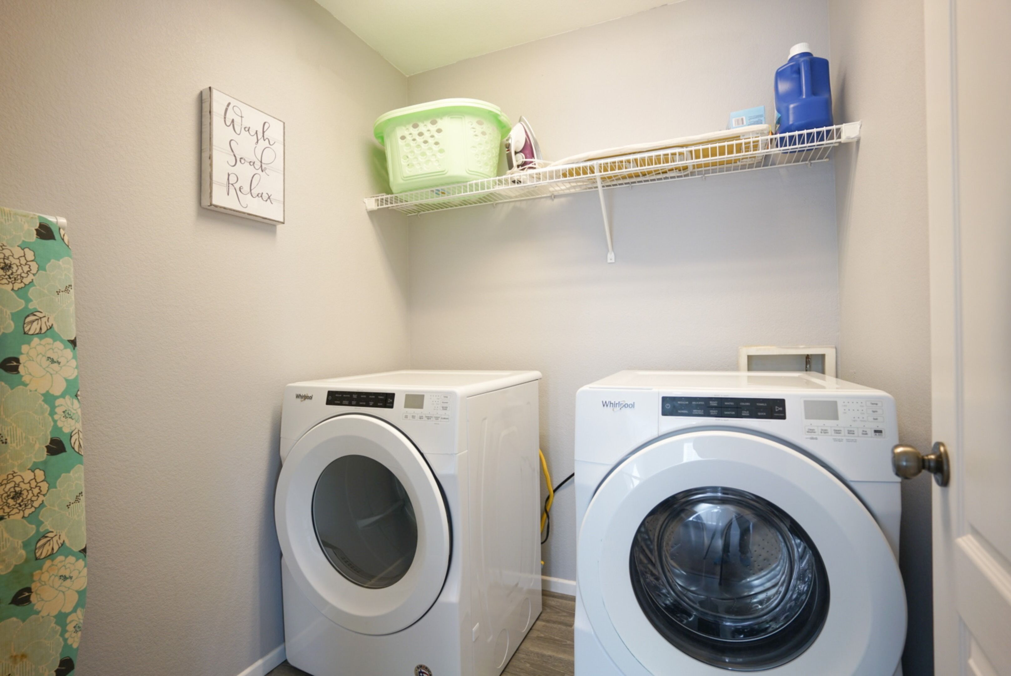 A laundry room with two front-loading washing machines, a shelf with laundry supplies, and a decorative wall sign.