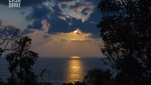 Tramonto sull'oceano con nuvole, palme e un logo nell'angolo.
