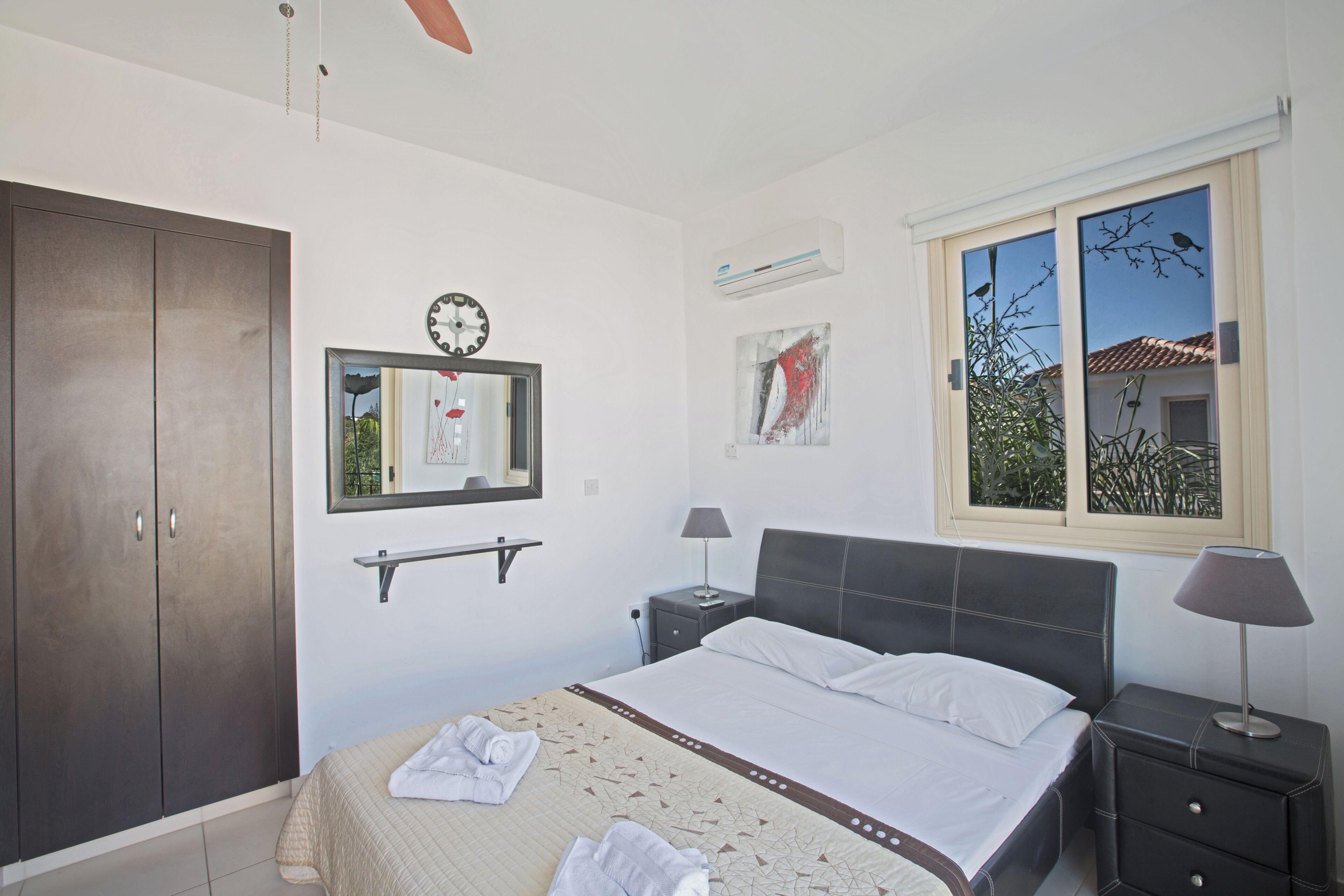A bedroom with a bed, a wardrobe, a bedside table with a lamp, a mirror, and a window with a view of trees.