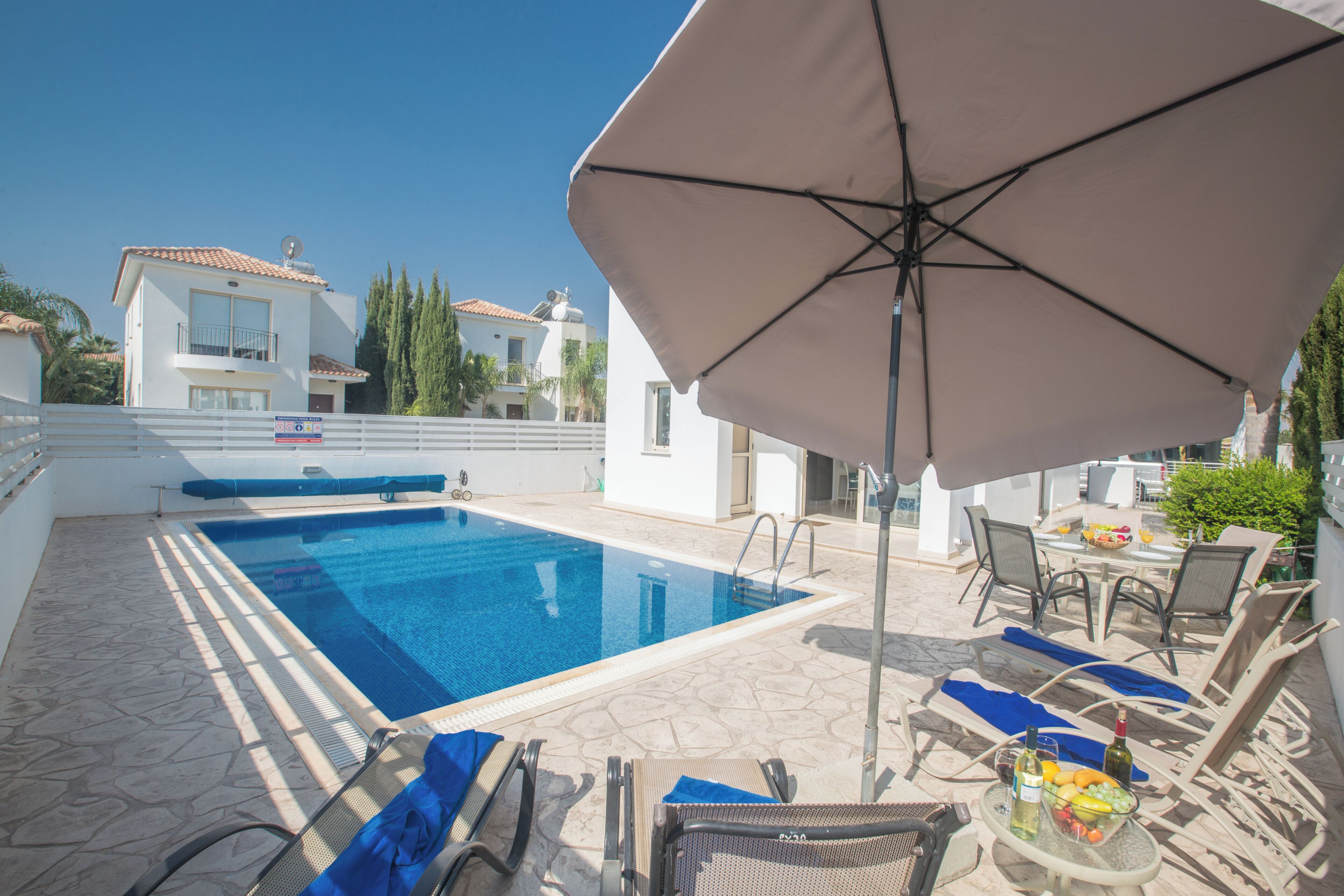 A poolside area with lounge chairs, an umbrella, and a table with fruits and a bottle.