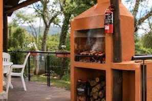 Een buitenoven van baksteen, met een barbecue, een brandblusser, een stoel en houtblokken in de buurt.