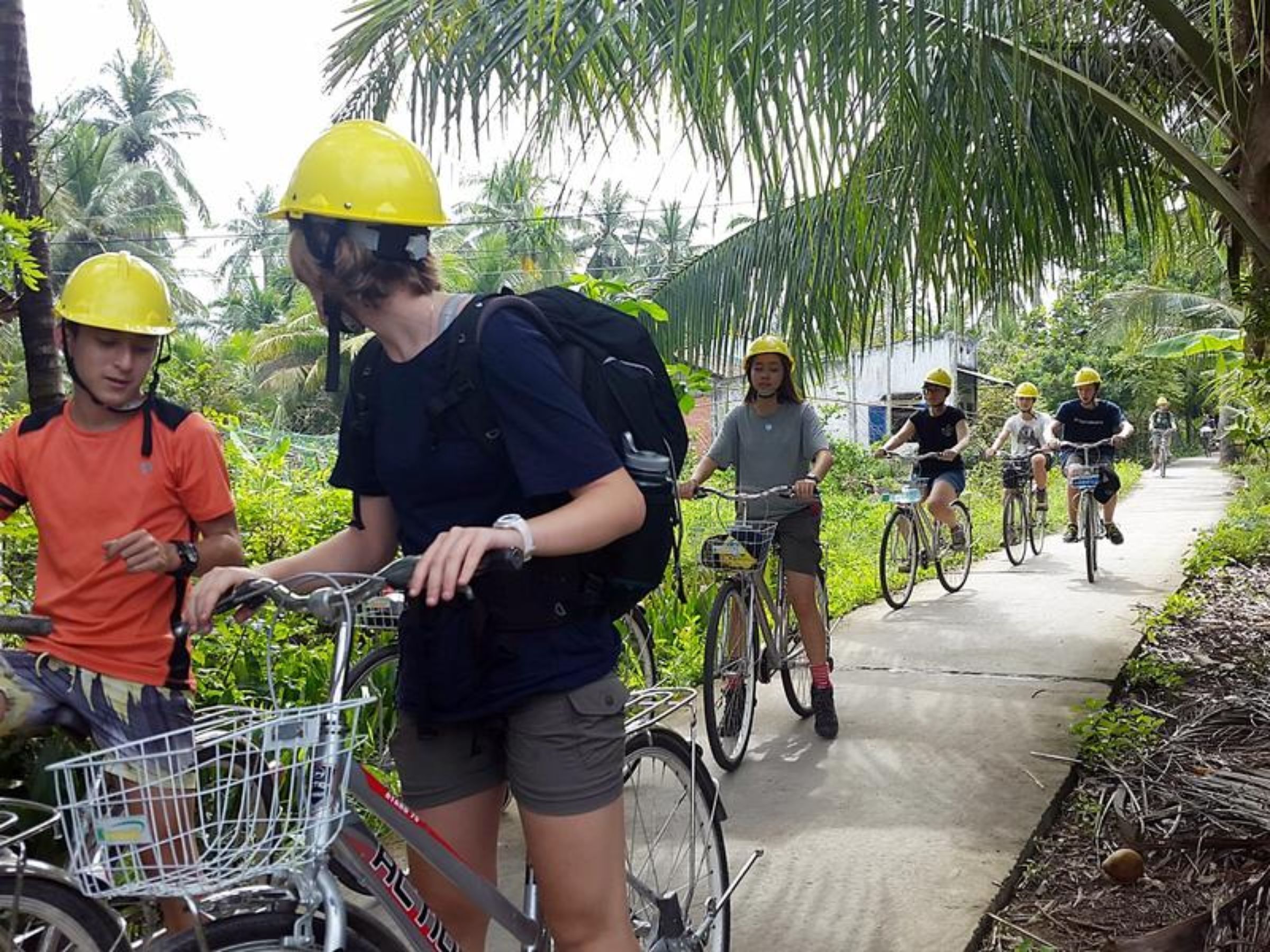 A group of people on bicycles, some wearing helmets, on a paved path surrounded by greenery.