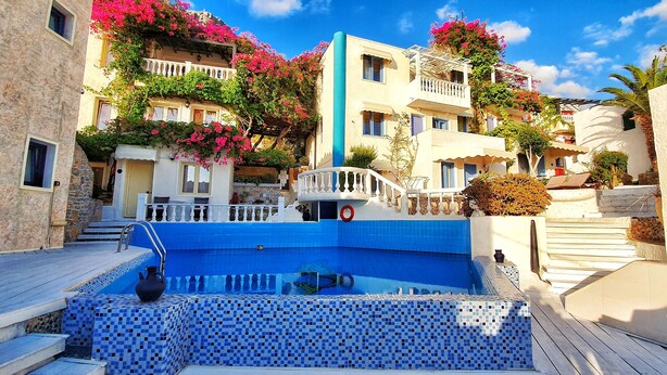 Un espace piscine doté d’un sol carrelé de bleu, entouré de bâtiments avec des balcons et des fleurs roses.