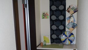 A narrow hallway with a door on the left, a cabinet with a fan and decorative items on the right, and a patterned wall hanging above. - Apartment A at Asahikawa City (Asahikawa)