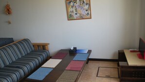 A living room with a sofa, a coffee table with placemats, a television, and a wall-mounted picture frame. - Apartment A at Asahikawa City (Asahikawa)