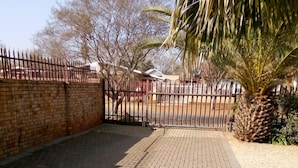 A paved courtyard with a brick wall, a metal gate, and a palm tree. - Casa Villa Guest House Carletonville - Suite 4 (Merafong City)